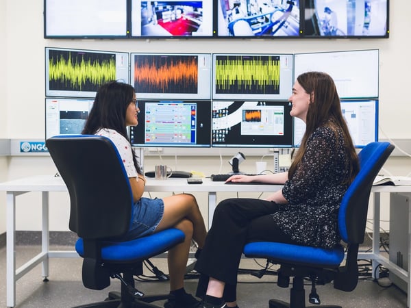 Two students in discussion with a wall of monitors displaying various data graphs